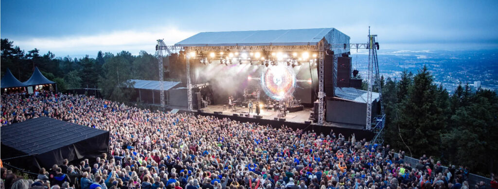 Stor utendørs musikkfestival i Oslo med tusenvis av mennesker foran en opplyst scene omgitt av trær og utsikt over byen.